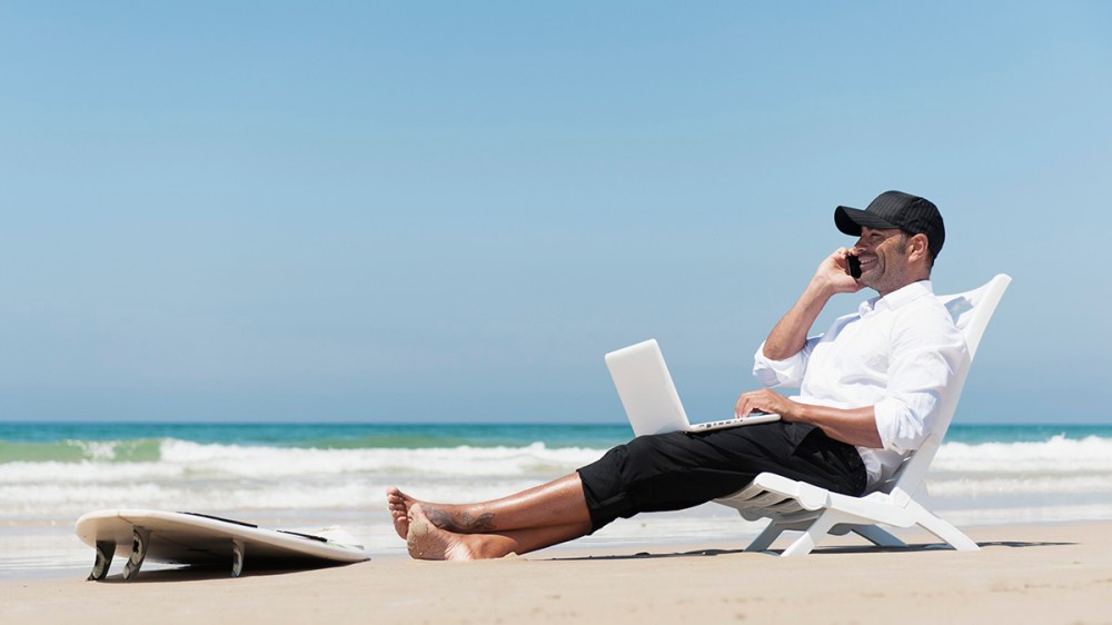 A Businessman Sits On A Beach Chair On The Beach Working On A Laptop Computer And Talking On The Phone With His Surfboard Sitting At His Feet; Tarifa Cadiz Andalusia Spain